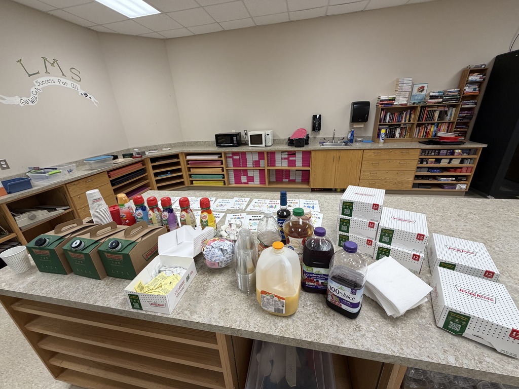 A room has a table filled with various items, including boxes, bottles, and a microwave. Behind the table, shelves hold books and items. "LMS" is written on the wall.