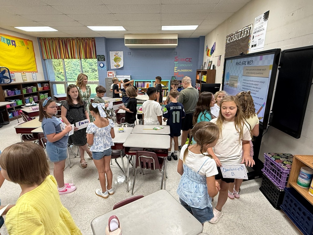 Elementary school students interacting and sharing name tags in a classroom setting during a "WHO ARE WE?" icebreaker activity. An interactive white board displays lesson prompts on the wall in the background alongside STEM educational displays.