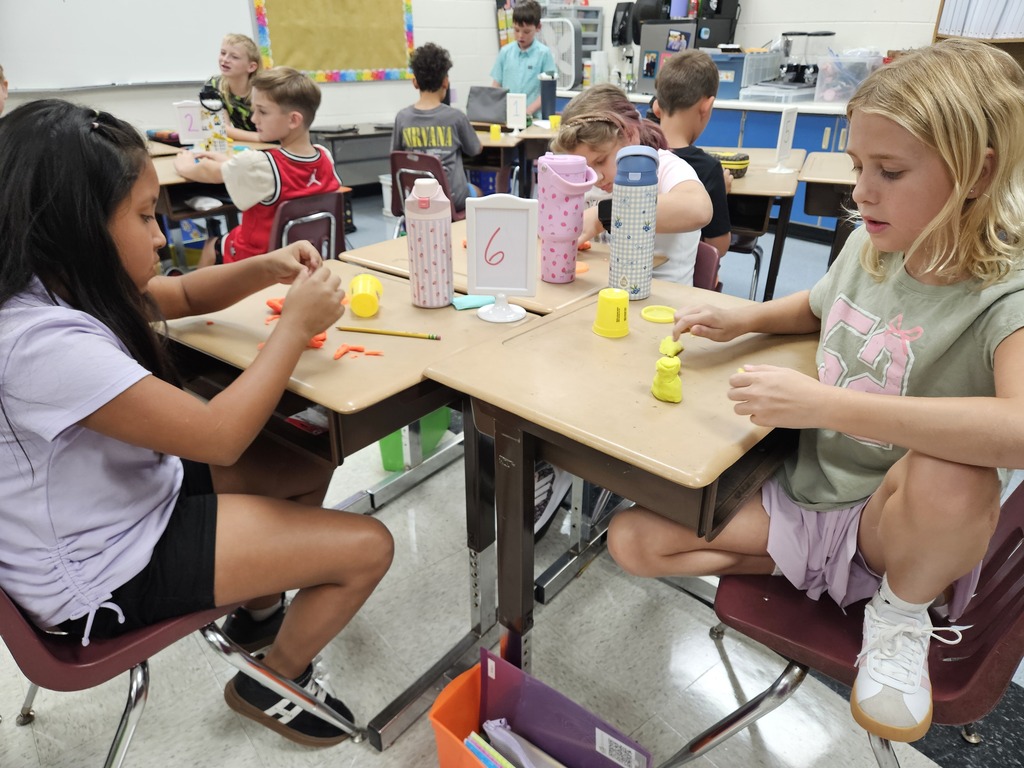 Elementary school students working at paired classroom desks using modeling compound during a hands-on activity. Water bottles and numbered station markers sit on the desks while other students work in the background.