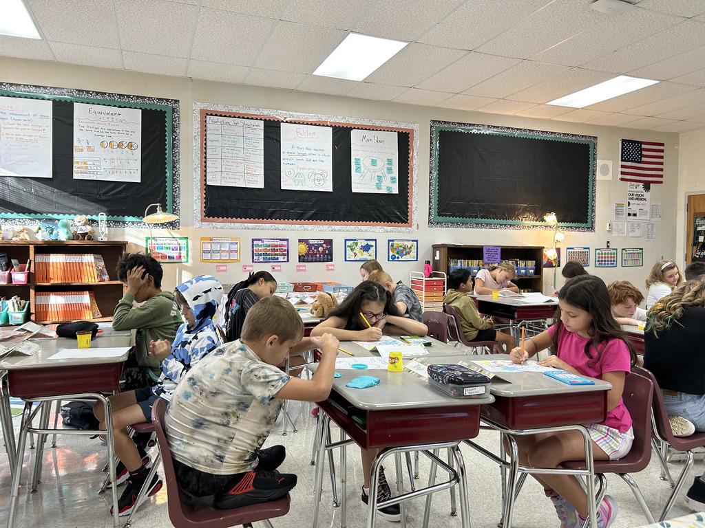 Elementary school students sitting at desks in a brightly lit classroom, focused on writing and working on desktop assignments. Educational posters displaying lessons on fractions, story structures, and main ideas line the classroom walls in the background.
