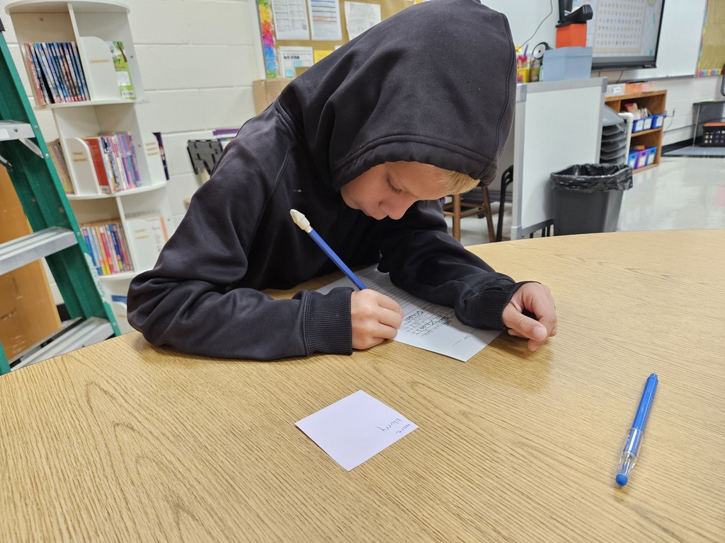 A student wearing a black hooded sweatshirt seated at a wooden desk, focused on writing on a paper with a blue pencil. A small purple sticky note and a blue pen sit nearby on the desk surface, with classroom bookshelves visible in the background.