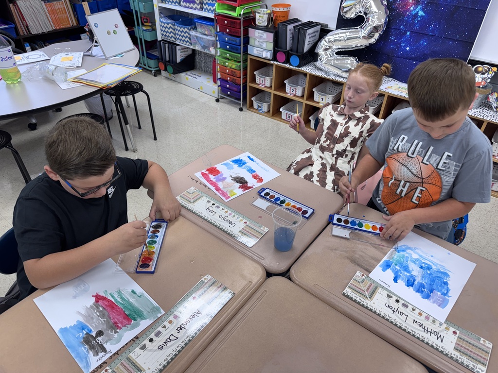 Three children in a classroom use paint palettes to mix colors at a table. Behind them is a classroom with shelves and a table.