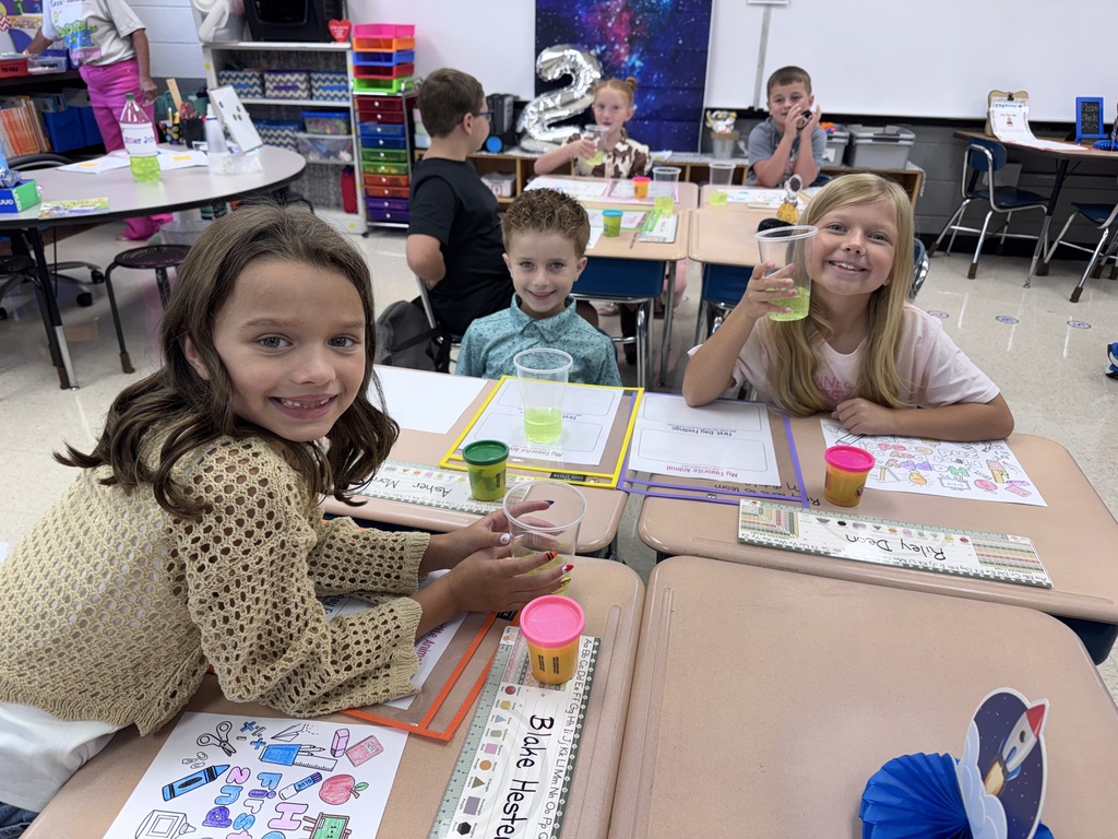 Children sitting at desks in a classroom, some holding cups, and a teacher's desk in the background.