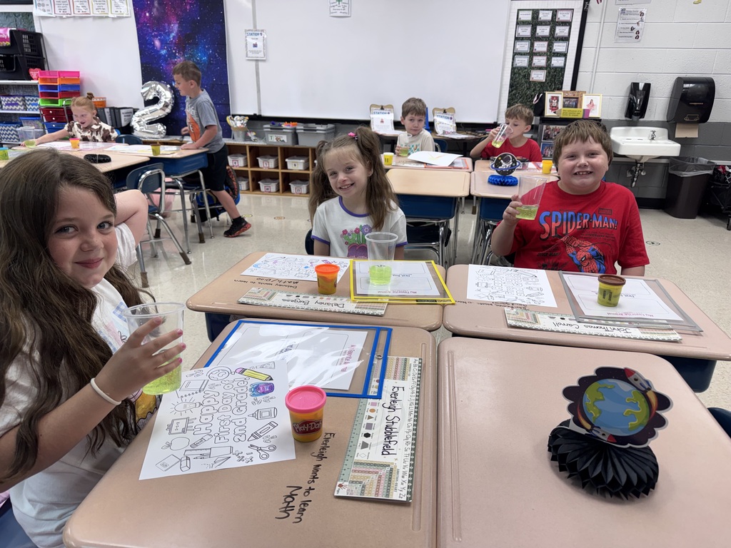 A classroom with children at desks. One child is holding a cup. A globe sits on a desk.