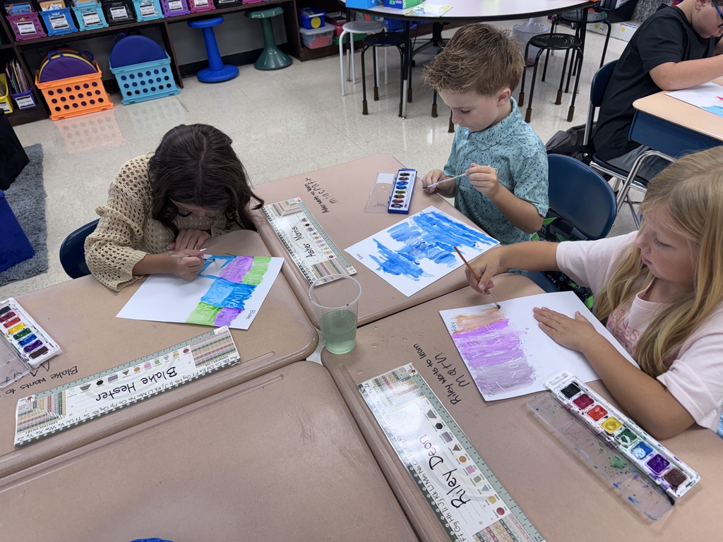 Three children at desks in a classroom, painting on paper with paintbrushes and paint palettes.