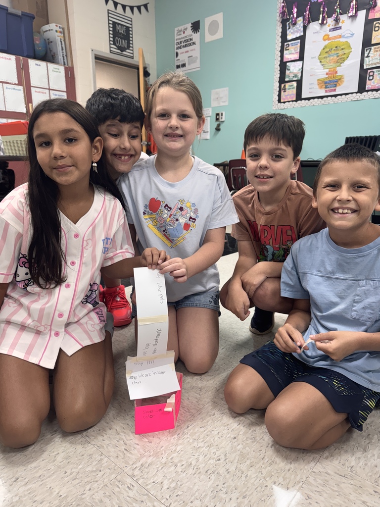 Five elementary school students kneeling together on a classroom tile floor, smiling while displaying a small paper tower structure with handwritten notes taped to it. Educational posters and classroom storage are visible in the background.