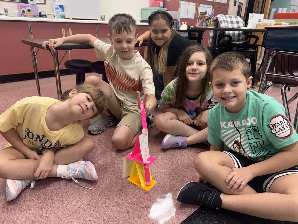 Five elementary school students sitting in a circle on the classroom floor, smiling next to a colorful paper tower structure they built together. Classroom tables and cabinets are visible in the background.