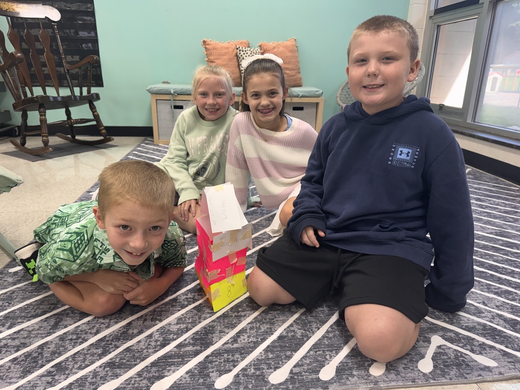 Four elementary school students gathered on a pattern rug in a classroom, posing next to a colorful paper tower activity featuring handwritten notes taped together.
