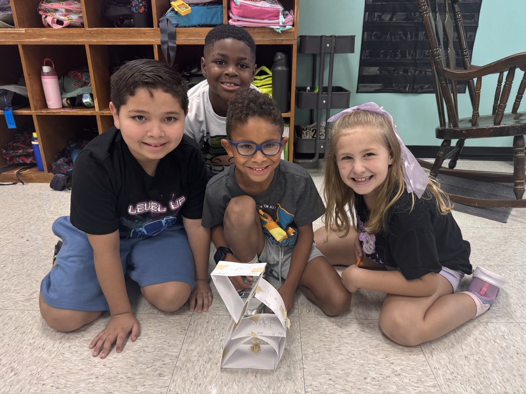 Four elementary school students smiling together on the classroom floor, posing behind a small tower structure crafted from paper and tape. Cubbies filled with backpacks and water bottles are visible in the background.