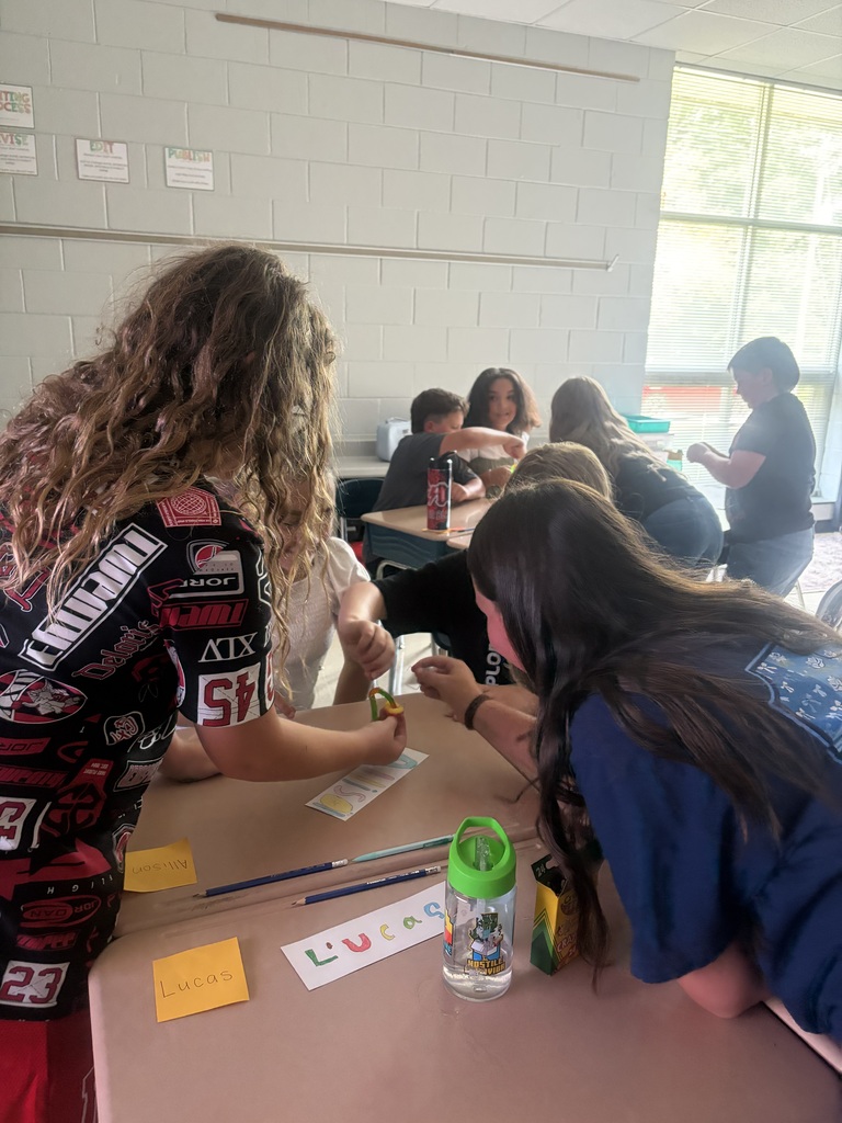 A group of people sit at a table, some engaged in activities. A woman with curly hair leans forward.