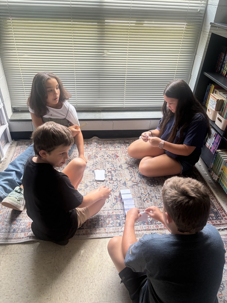 Four children sit on a rug, playing a card game in a room with a window.