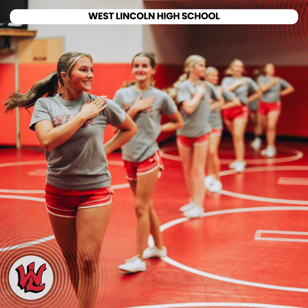 A group of girls practices cheerleading in a gymnasium. They wear matching uniforms and stand in a circle on a red mat.