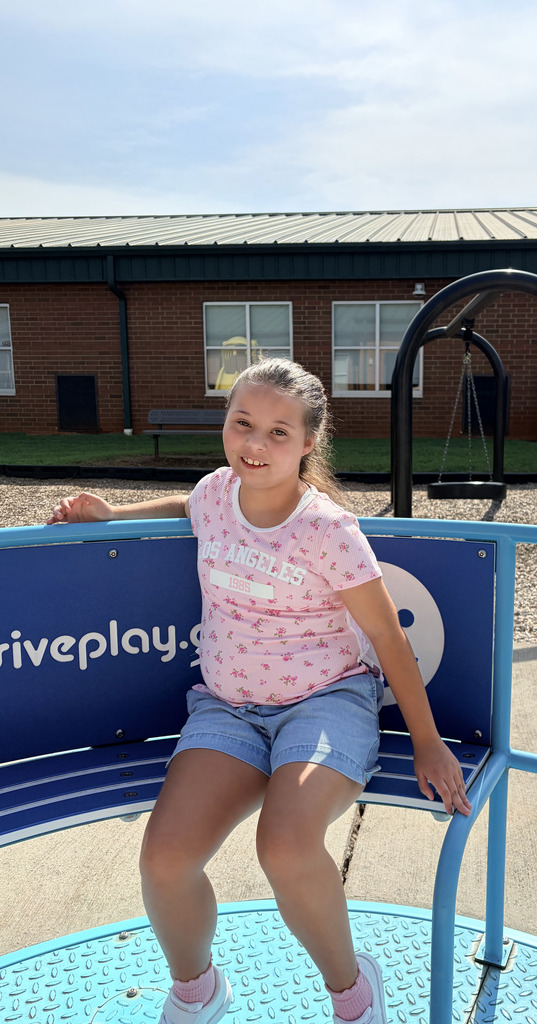 A girl sits on a blue bench at a playground, wearing a pink shirt and blue shorts. Behind her, a building and swing are visible.