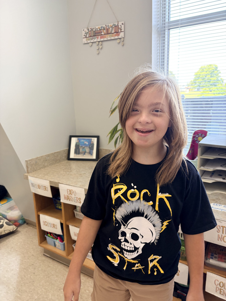A smiling girl with blonde hair stands in a classroom. She wears a black shirt with a skull design and yellow text.