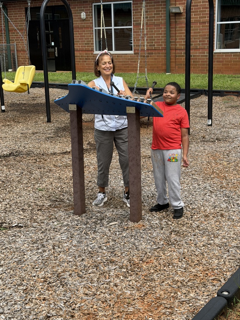 An adult and a child stand near a musical instrument in a playground with gravel ground.