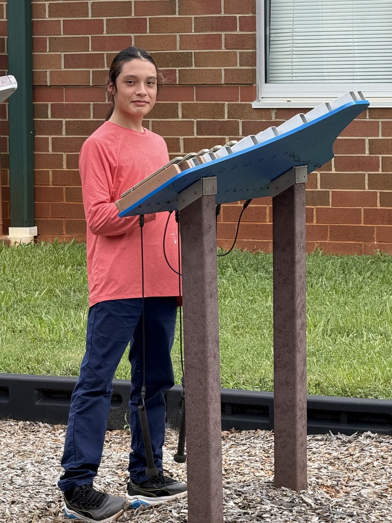 Person standing next to a blue music stand with an attached instrument. Brick wall and window in the background.