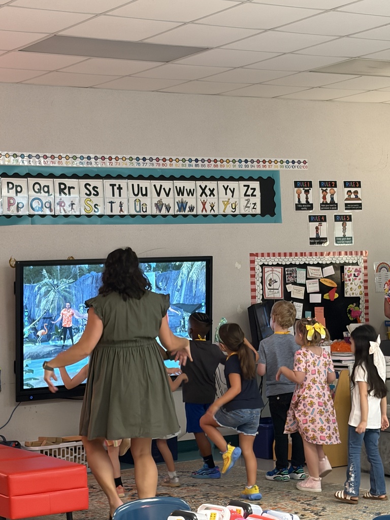 Woman leads children in front of screen; some wear sneakers. Alphabet displayed on wall. Bright lighting and indoor setting.