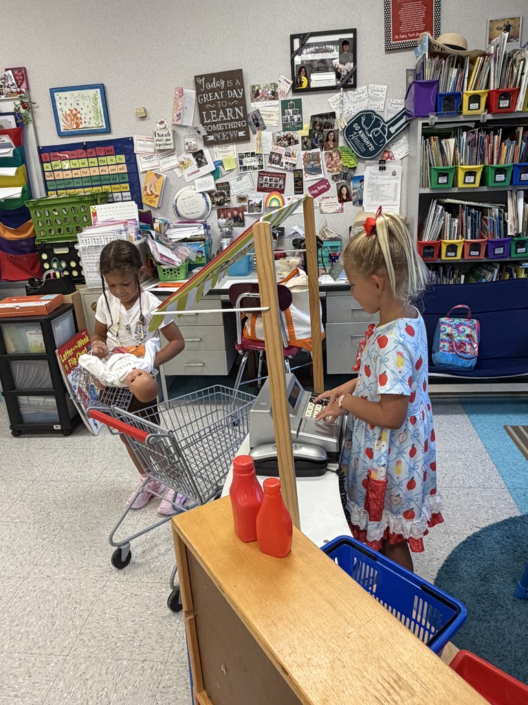 Two young girls are playing in a classroom. One girl pushes a shopping cart and the other stands behind a checkout counter.