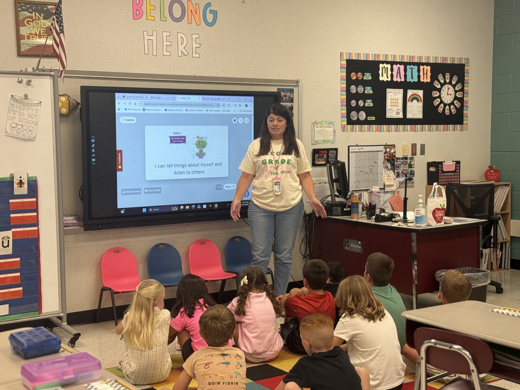 A woman stands in front of a screen in a classroom. Children sit on the floor and chairs.