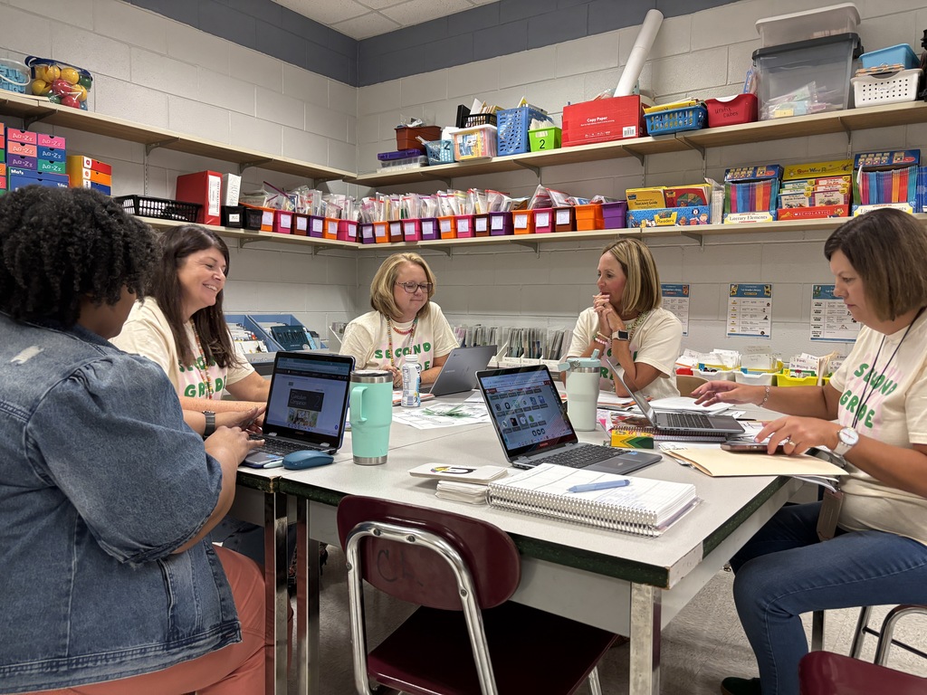 Five individuals are seated around a table in a classroom. Four are wearing light-colored shirts. They are focused on laptops and papers.