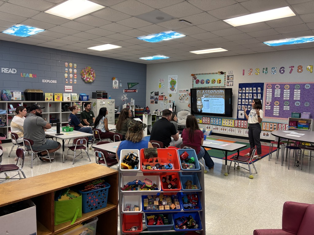 A classroom with multiple tables and chairs. A teacher stands in front of a projector screen. Students sit at tables and interact with each other.