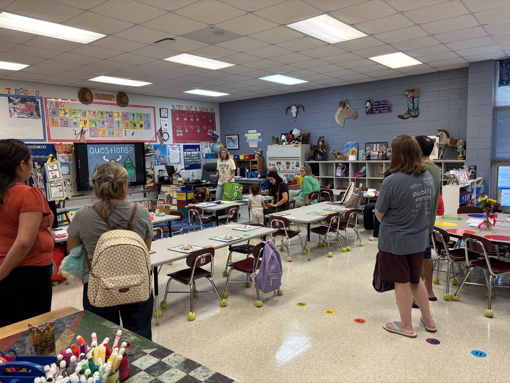A classroom with tables, chairs, and people. Some wear backpacks. Bright lights are on the ceiling.