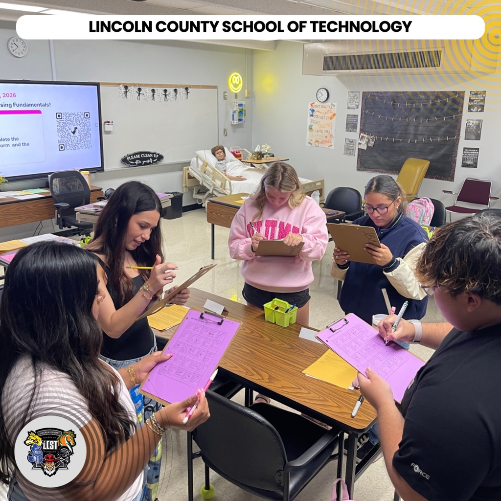 A classroom with students in pink and blue shirts. Four students are standing around a table writing on purple paper.