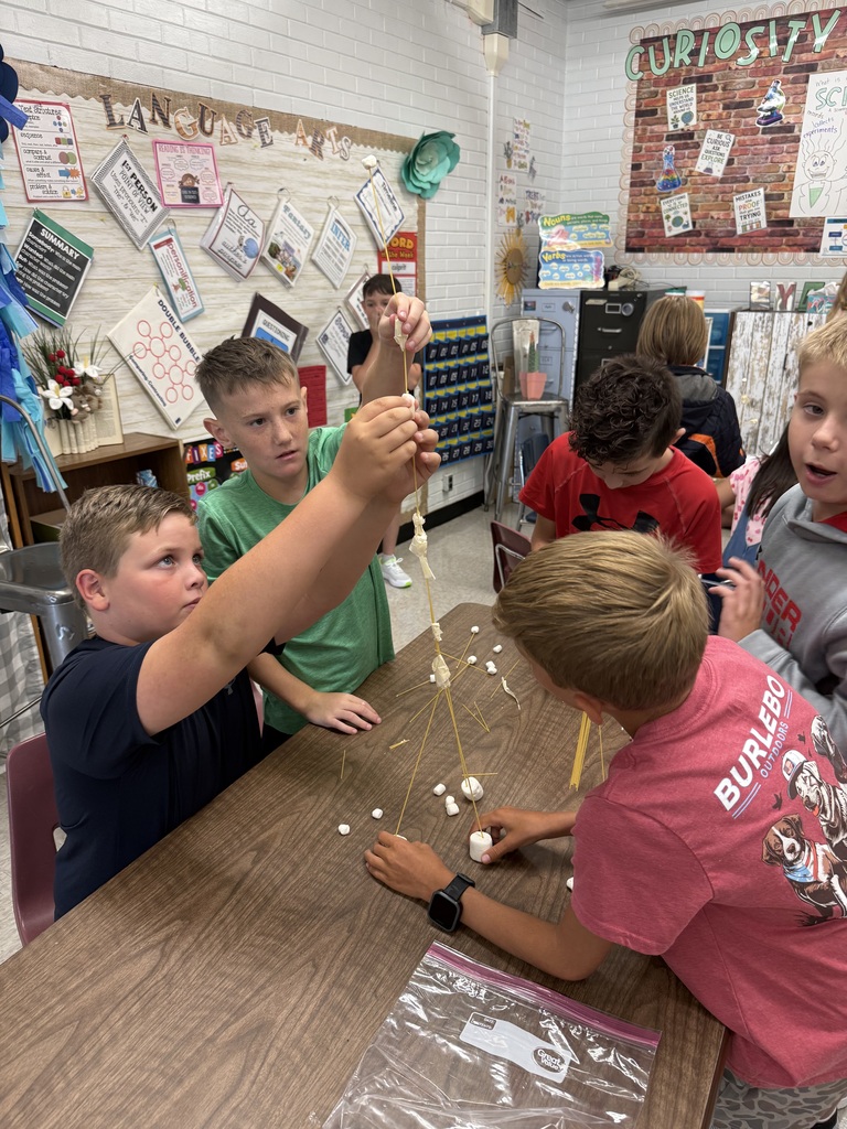 Several children sit around a table, working on a craft project. One child holds a string of pasta, and another holds a stick.