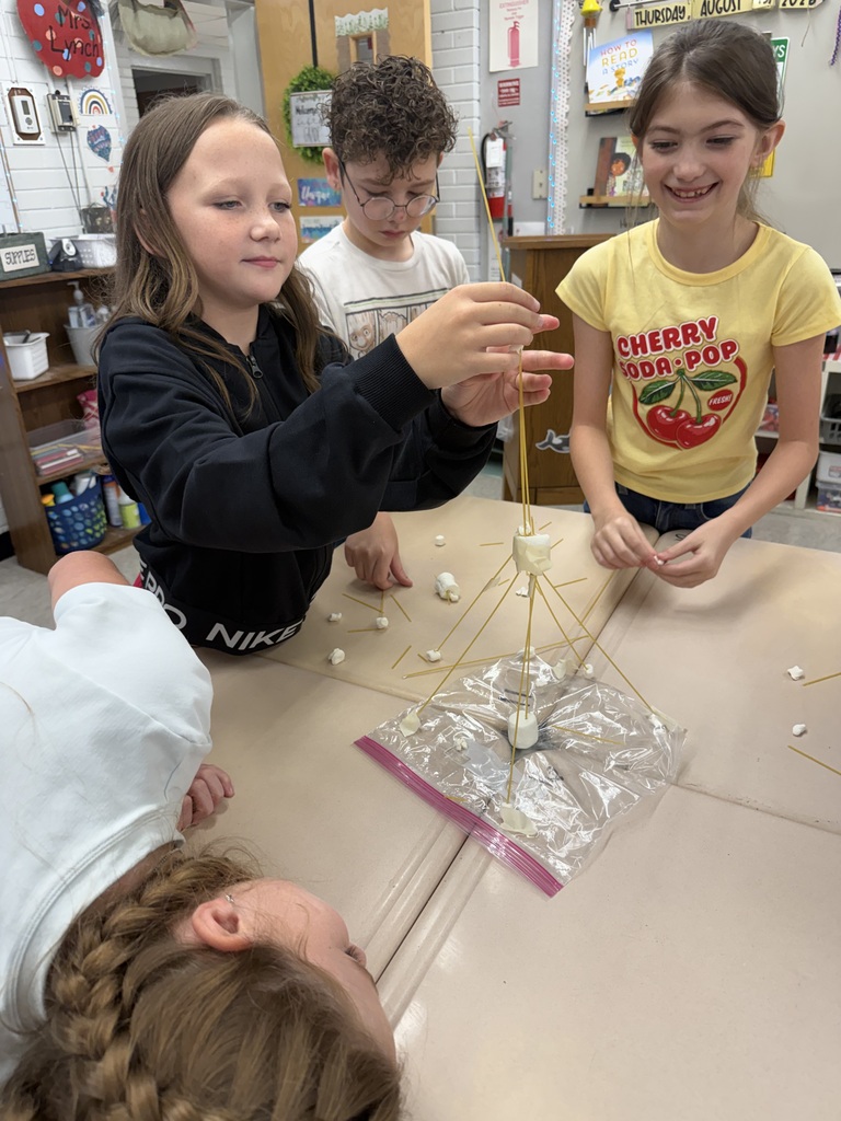 Four children collaborate on a table, constructing a model with sticks and plastic, one wearing a yellow shirt with "Cherry 100-POP" printed on it.