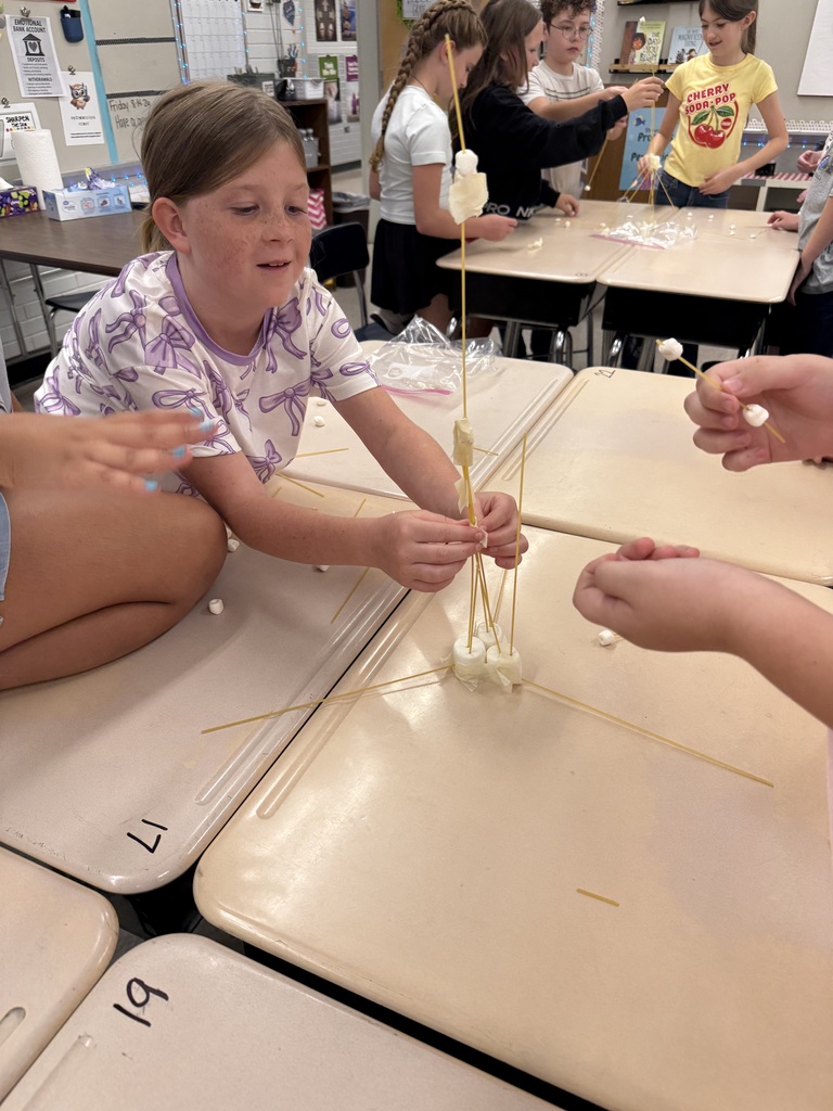 Children are sitting at desks, working together on a craft project involving marshmallows and sticks.