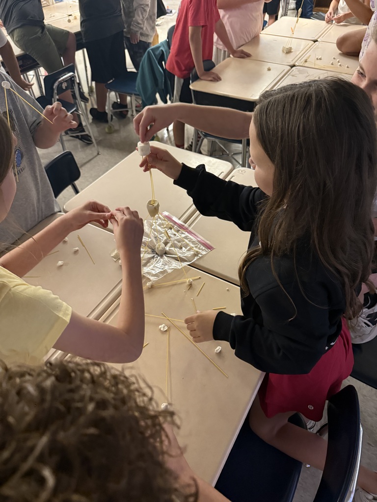 Children seated at tables, one holding a string and beads, another reaching for a similar string, with other kids nearby.