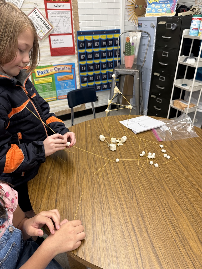 Two students build a structure with string and marshmallows on a table in a classroom.