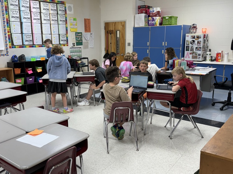 Students sit at individual desks arranged in a classroom setting, actively working on personal laptop computers while an educator works with students in the background.