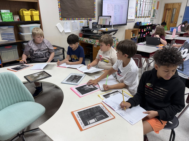 Five elementary students sit around a semi-circular table in a classroom, writing in notebooks alongside laminated worksheets with photos and text during a group lesson.
