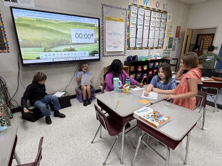 Elementary students collaborate in a classroom setting, working with pencils and papers at a cluster of desks while others sit near a large digital display board showing a timer screen.