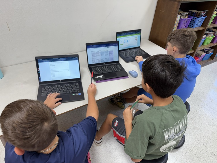 Three students sit side-by-side at a white table in a classroom, actively working on individual laptop computers open to educational programs and assignments.
