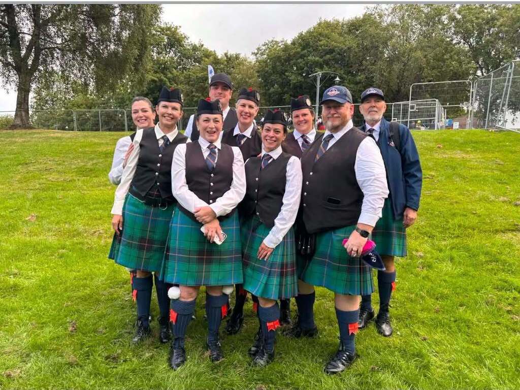 Group of individuals in kilts, posing for a photo on a grassy field with trees in the background.