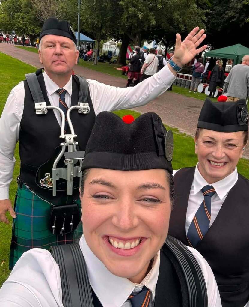 Three people in traditional Scottish attire smile in a park. One person waves. Trees and other people are in the background.
