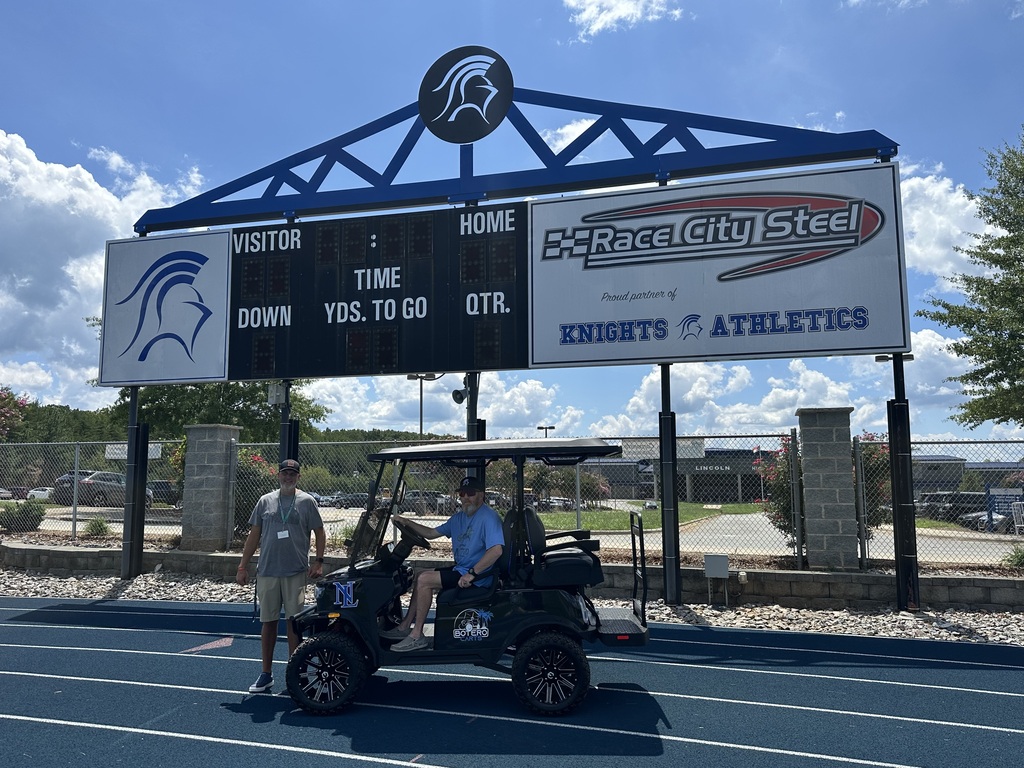 Two people, one standing and one seated, in a golf cart in front of a scoreboard.