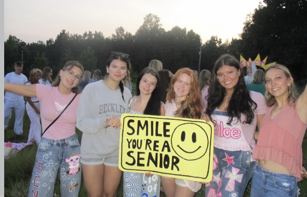 A group of young women hold a yellow sign that reads, "Smile, You're a Senior". They stand outdoors in front of trees.