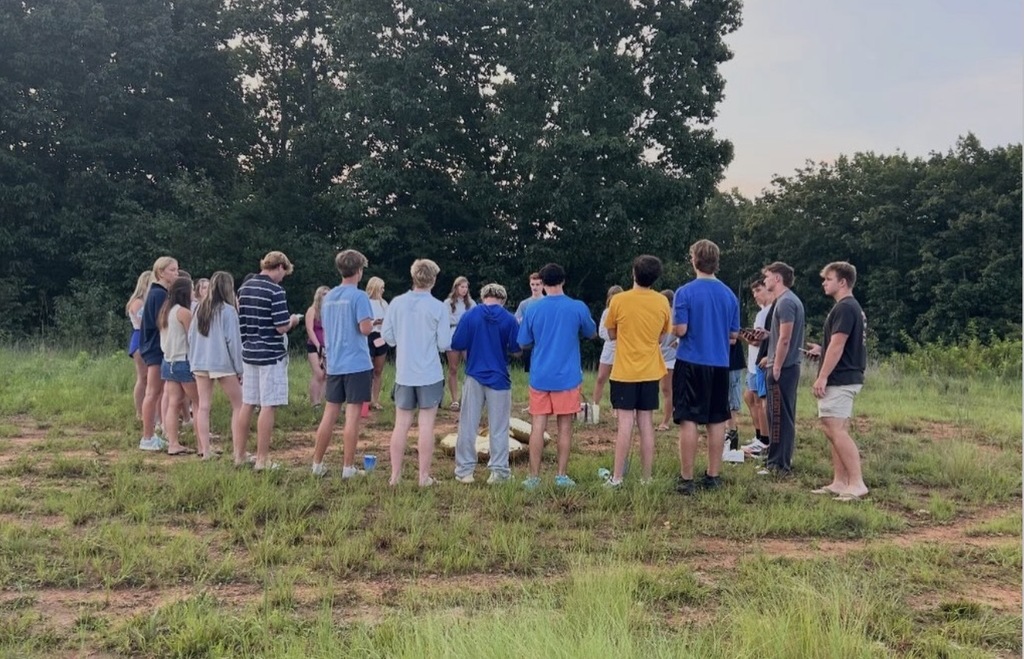 A group of people in casual attire standing in a circle on a grassy field, with trees in the background.