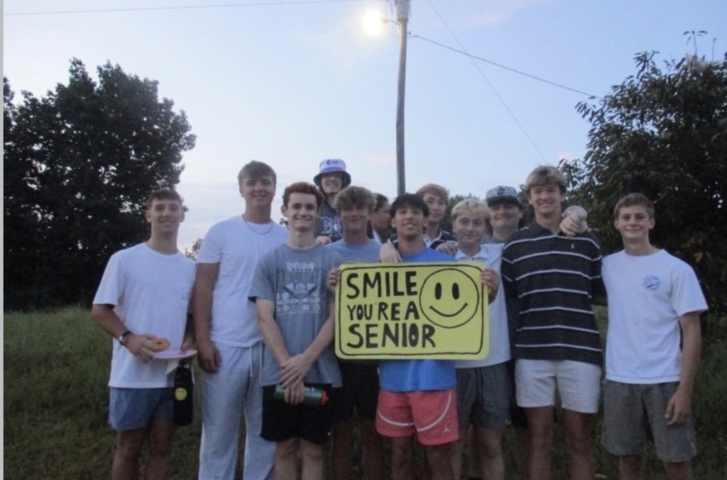 Group of young people in matching white shirts and shorts stand in a field with a sign reading "Smile, you're a senior".