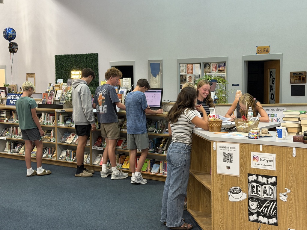 In a library, several students stand at a counter and at shelves, with some using computers.