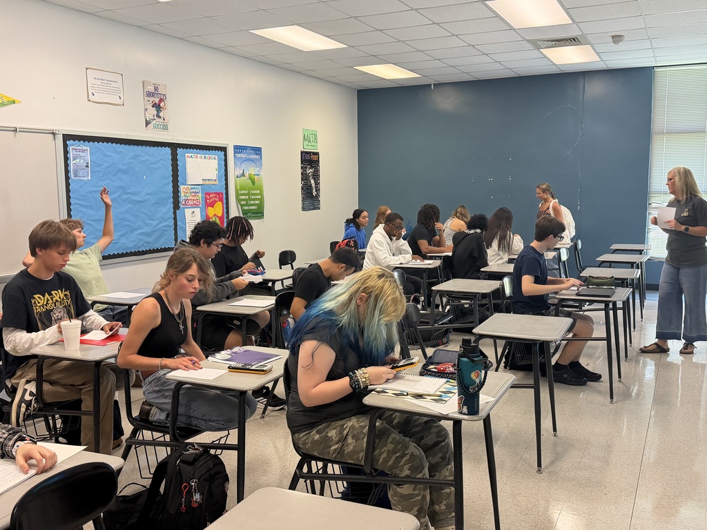 Classroom with students seated at desks. Teacher stands at the front of the room. Desks are arranged in rows. White walls and blue partition.