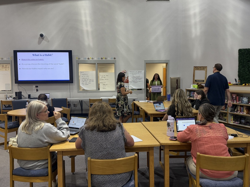 People in a classroom setting, seated at desks, with a woman standing in front of a screen.