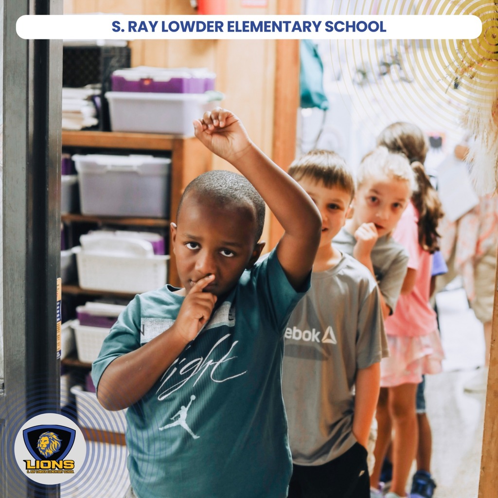 Several children stand in a school hallway. One child with raised arms and a hand to their mouth.