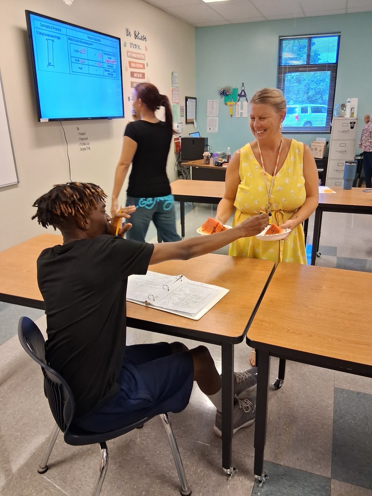 Two people in a classroom. One person hands another a plate of food. One person eats while seated.