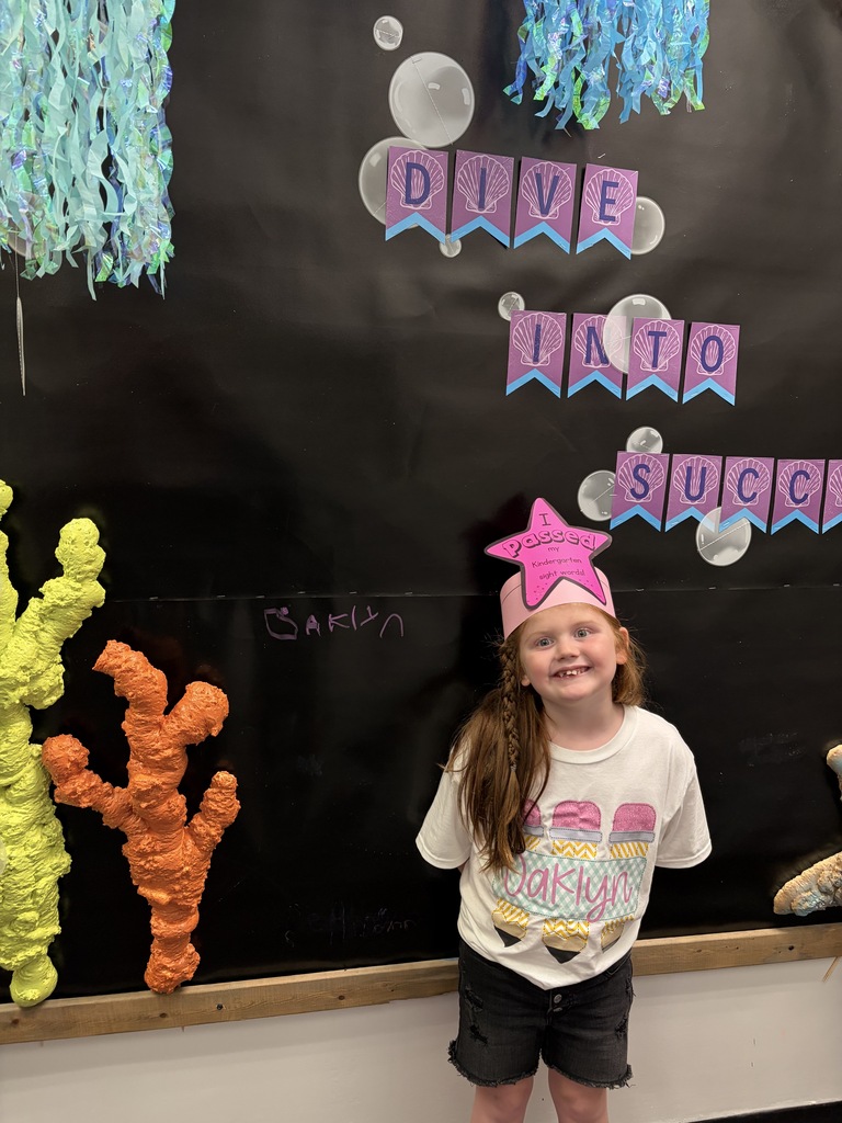 A child stands in front of a black bulletin board with a coral reef theme, wearing a pink star hat.