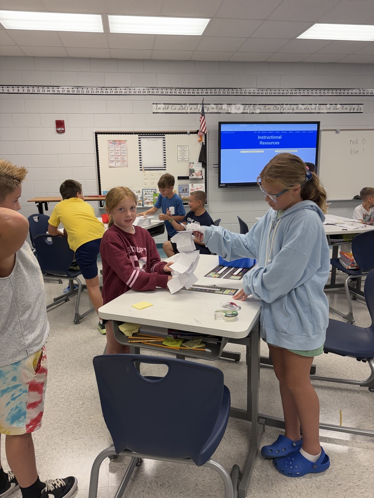 A classroom with students working at desks. One girl hands out papers while another girl holds them.