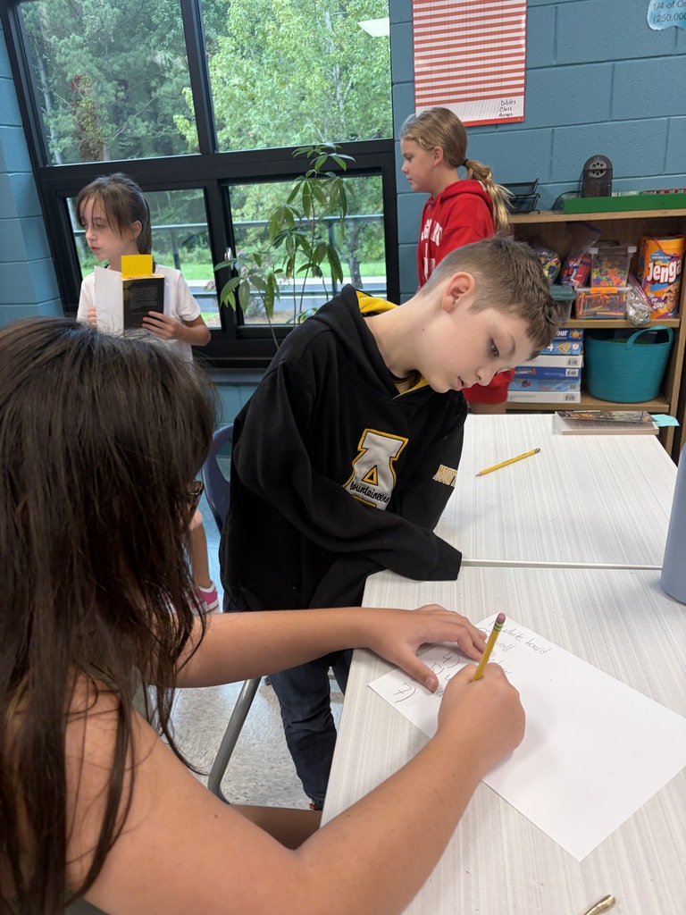 A boy and girl in a classroom writing on paper at a desk, with another student behind them.